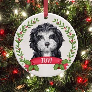 May include: A round white ceramic ornament featuring a black and white puppy illustration centred within a green holly leaf wreath with red berries. A red banner across the bottom displays the name JOVI in white text, suspended by a red ribbon.