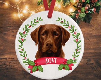 May include: A round white ceramic ornament featuring a brown Labrador retriever portrait centered within a green holly leaf wreath with red berries. A red ribbon banner across the bottom displays the name JOVI in white capital letters.