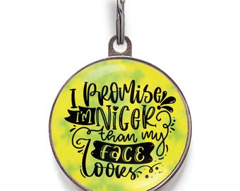 May include: A round metal pet ID tag with a yellow and green tie-dye background. The tag features black decorative lettering that reads, I PROMISE I'M NICER than my face LOOKS. A silver metal jump ring is attached to the top of the circular tag.