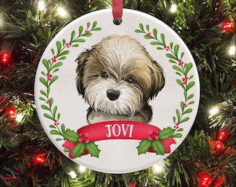May include: A round white ceramic ornament featuring a brown and white dog illustration centered within a green holly wreath with red berries. A red banner across the bottom displays the name JOVI in white text. A red ribbon hangs from the top.