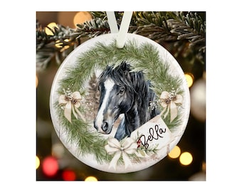 May include: A round white ceramic ornament featuring a black horse with a white blaze on its face, framed by a green pine wreath with cream-colored bows and pinecones. A small tag attached to the wreath displays the name Bella in black script.