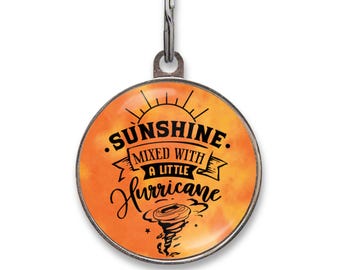 May include: A round metal pet ID tag with a silver-toned border and a split ring attachment. The center features an orange watercolor-style background with black text that reads Sunshine Mixed With A Little Hurricane and a small tornado graphic.