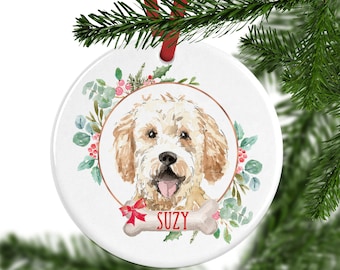 Op de afbeelding: Een rond wit keramieken ornament met een aquarelillustratie van een goudkleurige hond met een zwarte neus en open bek. De hond is omringd door groene bladeren en rode bessen, met daaronder een botvormige afbeelding met de naam SUZY.