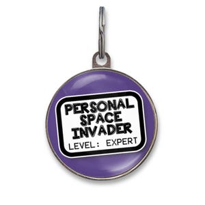 May include: A round purple pet ID tag with a metal attachment ring. The center features a white rectangular graphic with a black border containing the black text: PERSONAL SPACE INVADER LEVEL: EXPERT. The design has a smooth, glossy finish.
