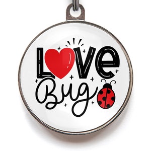 Puede incluir: Una etiqueta redonda de plata para mascotas con fondo blanco y texto negro que dice "Love Bug". También se muestra un corazón rojo y una mariquita roja con manchas negras en la etiqueta.