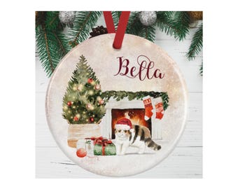 May include: A round ceramic Christmas ornament featuring a watercolor illustration of a cat wearing a Santa hat, a decorated Christmas tree, wrapped gifts, and a fireplace with stockings. The name Bella is printed in red script at the top.
