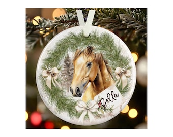 May include: A round white ceramic ornament featuring a brown horse head centered within a green pine wreath. The design includes three cream-colored bows, pinecones, and a white gift tag with the name Bella written in black cursive script.