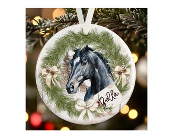 May include: A round white ceramic ornament featuring a black horse head illustration centered within a green pine wreath. The design includes three cream-colored bows and a small tag with the name Bella written in black cursive script text.