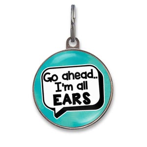 May include: A round, teal-colored pet ID tag featuring a white speech bubble graphic with the black text "Go ahead.. I'm all EARS". The circular metal tag includes a silver-toned metal jump ring at the top for attachment to a collar or accessory.