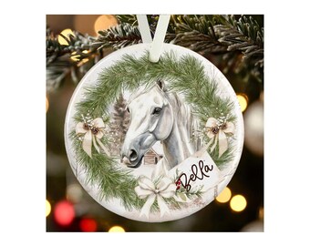 May include: A round white ceramic ornament featuring a printed illustration of a white horse head centered within a green pine wreath. The wreath is decorated with three cream-colored bows and a small tag with the name Bella written in black script.