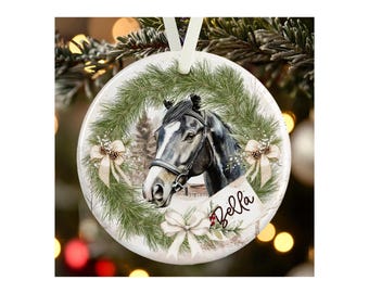 May include: A round white ceramic ornament featuring a dark horse with a white blaze on its face, framed by a green pine wreath with cream-colored bows and pinecones. The name Bella is written in black script on a small tag attached to the wreath.