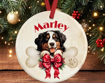 Op de afbeelding: Een rond keramieken ornament met een illustratie van een Berner Sennenhond en de naam Marley in het rood erboven. De hond rust zijn poten op een groot bot, versierd met een feestelijke rode strik, groene dennentakken en rode bessen.