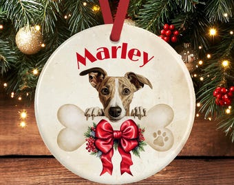 Op de afbeelding: Een ronde keramische kerstornament met een bruin-witte hond die zijn poten op een groot bot laat rusten. De naam Marley is in het rood boven de hond gedrukt. Een grote rode strik met groene hulst en rode bessen bevindt zich onder het bot, en een rood lint hangt aan de bovenkant.