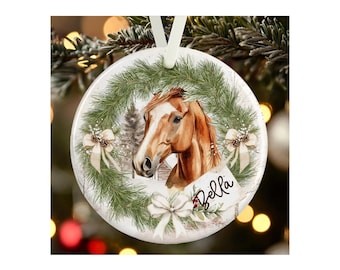 May include: A round white ceramic ornament featuring a brown horse head centered within a green pine wreath. The wreath is decorated with three cream-colored bows and small pinecones. The name Bella is written in black script on a small white tag.