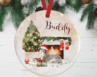 May include: A round ceramic Christmas ornament featuring a watercolor illustration of a grey dog wearing a Santa hat, sitting by a decorated Christmas tree and a fireplace with two stockings. The name Buddy is printed in dark red script above.