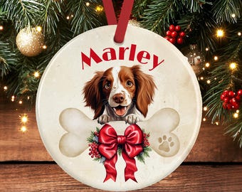 Op de afbeelding: Een ronde keramische kerstornament met een illustratie van een bruin-witte spaniël boven een groot bot. De naam Marley staat in rode letters boven de hond. Een felrode strik met groene hulstaccenten bevindt zich onder het bot.
