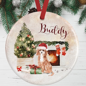 Könnte beinhalten: Ein runder Weihnachtsanhänger aus Keramik mit einem Aquarellbild eines braunen Hundes mit Weihnachtsmütze, der vor einem Kamin mit geschmücktem Baum und Geschenken sitzt. Der Name Buddy ist oben in dunkelroter Schreibschrift gedruckt.