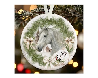 May include: A round white ceramic ornament featuring a gray horse centered within a green pine wreath. The design includes three cream-colored bows, small pinecones, and a white tag with the name Bella written in black cursive script.
