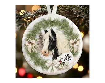 May include: A round white ceramic ornament featuring a black and white horse portrait framed by a green pine wreath. The design includes three cream-colored bows, pinecones, and a white gift tag with the name Bella written in black script.