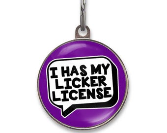May include: A round purple pet ID tag featuring a white speech bubble graphic with black text that reads I HAS MY LICKER LICENSE. The tag includes a silver metal border and a matching silver split ring attachment at the top for secure collar placement.