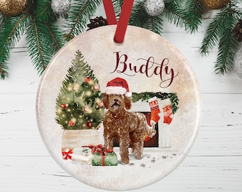 May include: A round ceramic Christmas ornament featuring a brown doodle dog wearing a red Santa hat. The illustration includes a decorated Christmas tree, a fireplace with two red stockings, and wrapped gifts. The name Buddy is printed in red.