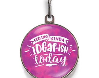 May include: A round metal pet ID tag with a glossy finish featuring a vibrant magenta and purple tie-dye background. The white text printed on the front reads, "FEELING KINDA IDGAF-ish TODAY," accompanied by a small metal split ring for attachment.