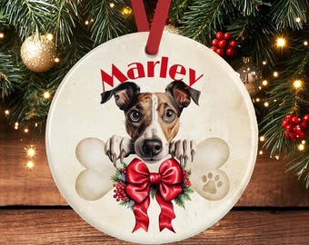 May include: A round ceramic Christmas ornament featuring a brown and white dog with the name Marley printed in red above its head. The dog rests its paws on a large bone graphic, accented by a bright red bow with green pine and red berries.
