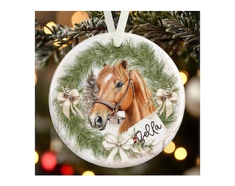 May include: A round white ceramic ornament featuring a brown horse head centered within a green pine wreath. The wreath is decorated with three cream-colored bows and pinecones. The name Bella is printed in black script on a white tag.