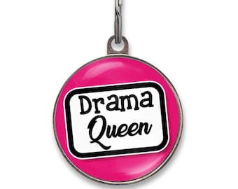 May include: A round metal pet ID tag with a bright pink background. In the centre, a white rectangular label with a black border features the text "Drama Queen" in a bold, black, stylised font. A silver metal jump ring is attached to the top.
