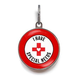 May include: A round, red and white pet tag with the text "I HAVE SPECIAL NEEDS". The tag features a red cross with a white heart in the center. A silver metal loop is at the top for attachment.