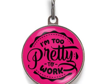 May include: A round, hot pink metal pet ID tag with a silver-toned metal border and a matching silver split ring at the top. The tag features black text that reads I'M TOO pretty TO WORK, arranged in a circular design with a scalloped black border.