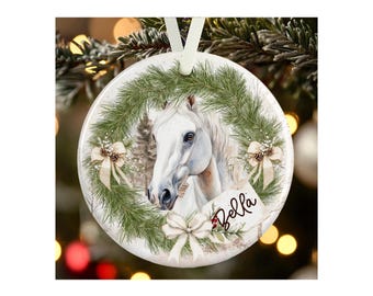 May include: A round white ceramic ornament featuring a printed illustration of a white horse framed by a green pine wreath with cream-colored bows and pinecones. A small rectangular tag attached to the wreath displays the name Bella in script.