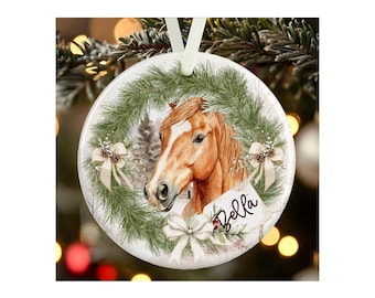 May include: A round white ceramic ornament featuring a brown horse illustration centered within a green pine wreath. The wreath is decorated with white bows and pinecones. A white tag with the name Bella written in black script is attached.
