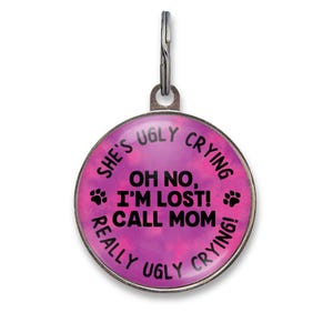 May include: A round metal pet ID tag with a pink and purple tie-dye background. The text reads: SHE'S UGLY CRYING, OH NO, I'M LOST! CALL MOM, REALLY UGLY CRYING! Two black paw prints are on the sides. A silver metal jump ring is attached to the top.