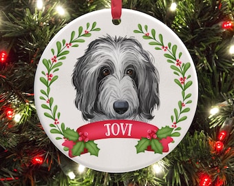 May include: A round white ceramic ornament featuring a detailed illustration of a grey and white shaggy dog. A red ribbon banner across the bottom displays the name JOVI in white capital letters, surrounded by a green holly and berry wreath.