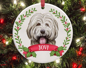 Op de afbeelding: Een rond wit keramieken ornament met een langharige hond met een roze tong, omlijst door een groene hulstkrans met rode bessen. Een rood lintbanner onderaan toont de naam JOVI in witte letters, inclusief een rood ophanglint.