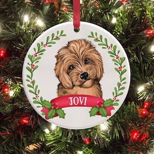 Puede incluir: Un adorno navideño redondo de cerámica blanca con una ilustración detallada de un cachorro marrón esponjoso. El cachorro está enmarcado por una corona de muérdago verde con bayas rojas. Una cinta roja en la base muestra el nombre JOVI en blanco.