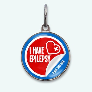Op de afbeelding: Een ronde, zilverkleurige medische waarschuwingshanger met een rood en wit ontwerp. De hanger heeft de tekst "I HAVE EPILEPSY" en een hart met een kruis. De rechterbenedenhoek heeft een sticker met "PLEASE TURN OVER".