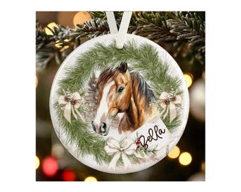 May include: A round white ceramic ornament featuring a brown horse head illustration centered within a green pine wreath. The design includes three cream-colored bows and a white tag with the name Bella written in black cursive script.