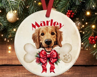 Puede incluir: Un adorno redondo de cerámica con la ilustración de un golden retriever y el nombre Marley impreso en rojo arriba. El perro se sitúa tras un gráfico de hueso blanco, decorado con un lazo rojo brillante, ramas de pino verdes y bayas rojas.