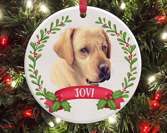 May include: A round white ceramic Christmas ornament featuring a yellow Labrador retriever illustration centered within a green holly wreath. A red ribbon banner across the bottom displays the name JOVI in white capital letters. A red ribbon loop hangs.