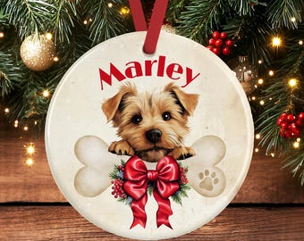May include: A round ceramic ornament featuring a brown puppy resting its paws on a large bone. The name Marley is printed in red above the puppy. A red ribbon bow with green holly leaves and red berries is centered below the bone, and a paw print is visible on the right side of the bone.