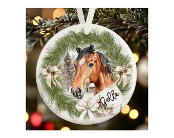 May include: A round white ceramic ornament featuring a brown horse head centered within a green pine wreath. The wreath is decorated with three cream-colored bows and pinecones. A white tag with the name Bella written in black script is attached.
