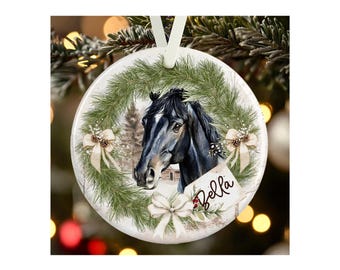 May include: A round white ceramic ornament featuring a black horse head illustration centered within a green pine wreath. The design includes three cream-colored bows and a small rectangular tag with the name Bella written in black script.