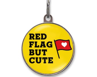 May include: A round yellow pet ID tag with a silver metal border and a split ring attachment. The tag features the black text RED FLAG BUT CUTE and a graphic of a red flag with a white heart in the center, printed on a bright yellow background.