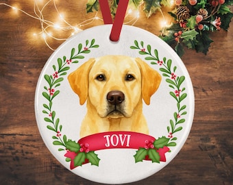 May include: A round white ceramic ornament featuring a yellow Labrador retriever illustration centered within a green holly wreath with red berries. A red ribbon banner across the bottom displays the name JOVI in white capital letters.