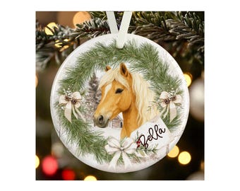 May include: A round white ceramic ornament featuring a golden horse head centered within a green pine wreath. The design includes three white bows, pinecones, and a small tag with the name Bella written in black script, suspended by a white ribbon.
