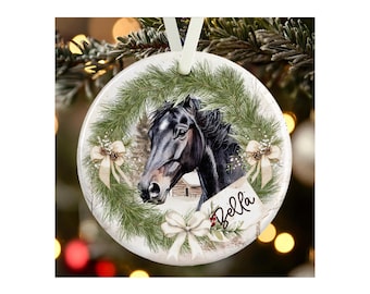 May include: A round white ceramic ornament featuring a black horse head illustration centered within a green pine wreath. The wreath is decorated with three cream-colored bows and a small tag with the name Bella written in black script.