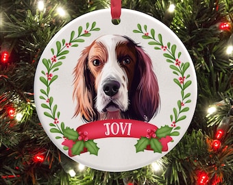 Op de afbeelding: Een ronde witte keramische kerstornament met een gedetailleerde illustratie van een bruin-witte hond. De hond is omringd door een groene hulstkrans met rode bessen. Een rode banner onderaan toont de naam JOVI in witte letters.