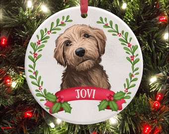 May include: A round white ceramic ornament featuring a brown dog illustration centered within a green holly wreath with red berries. A red ribbon banner across the bottom displays the name JOVI in white capital letters. A red ribbon loop hangs.